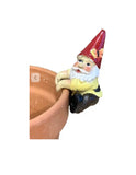 A small garden gnome pot sitter figurine with a red hat, yellow shirt, white beard, and black pants, shown leaning on the rim of a terracotta planter against a plain white background.